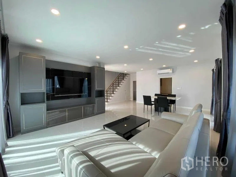 Detached House for rent in Don Mueang, Bangkok - Open-plan living and dining area with TV wall unit, staircase and glossy flooring.