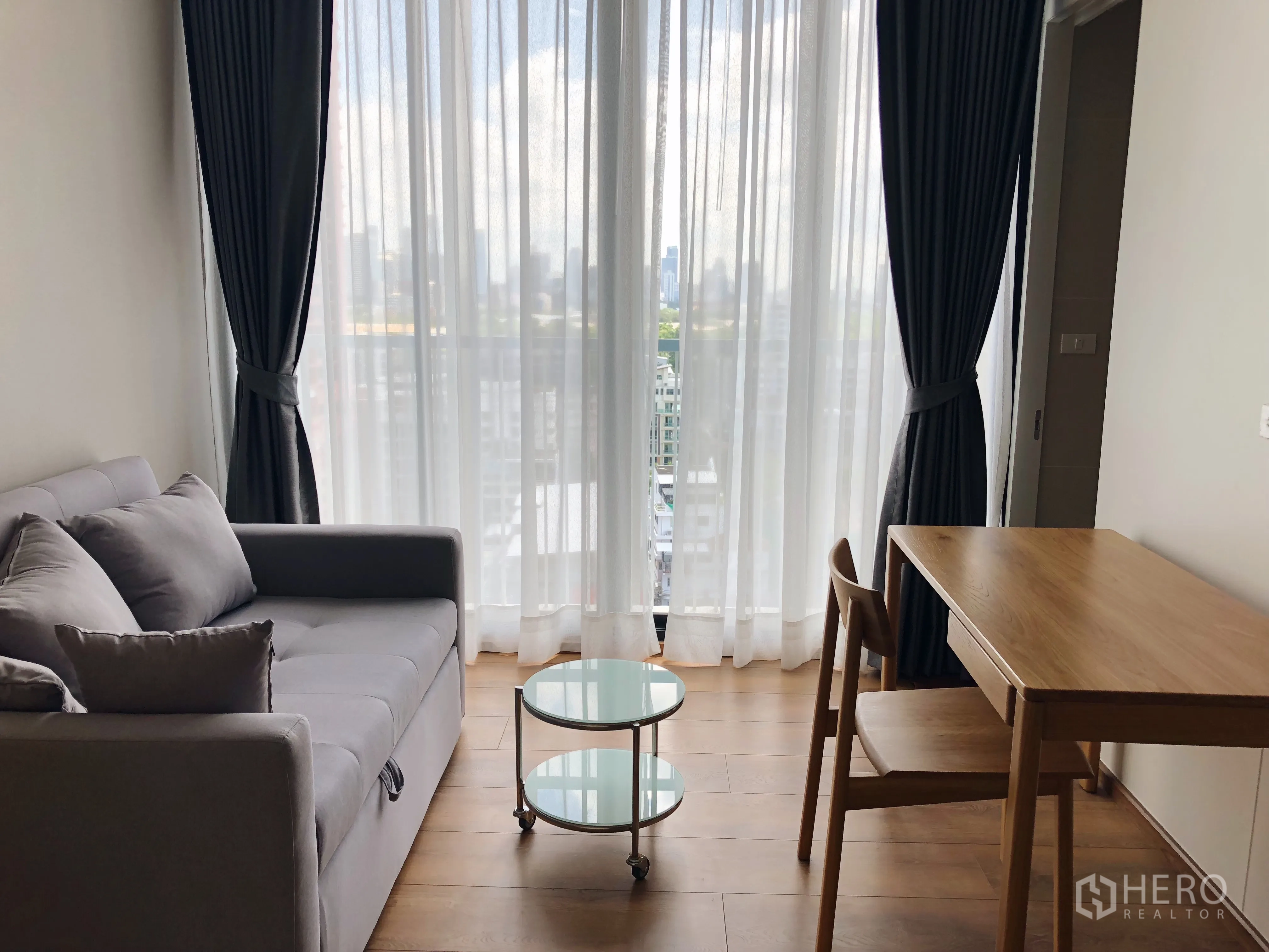 Condominium for rent in Khlong Toei, Bangkok - Compact lounge with gray sofa and wooden desk facing full-height windows and skyline outlook.