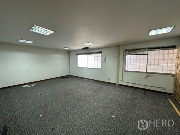 Warehouse for rent in Huai Khwang, Bangkok - Empty carpeted office room with fluorescent lighting and windows.