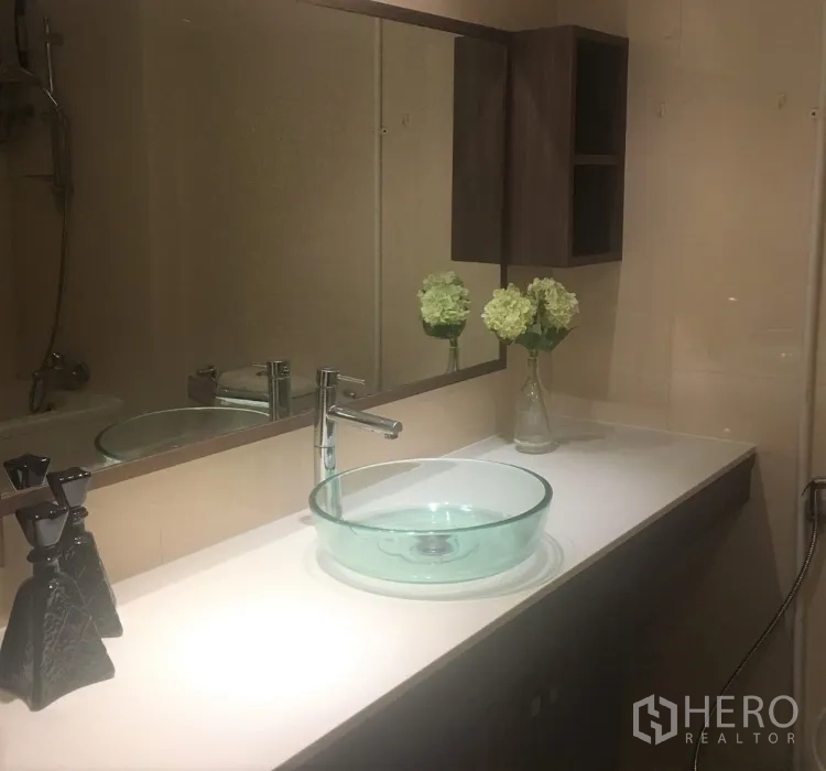 Condominium for rent in Watthana, Bangkok - Modern bathroom vanity with a glass vessel sink, long countertop, mirror, and wall cabinet.