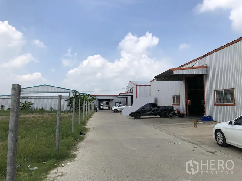 Warehouse for rent in Nonthaburi - Concrete access road between warehouse buildings with adjacent grassy plot and perimeter posts.