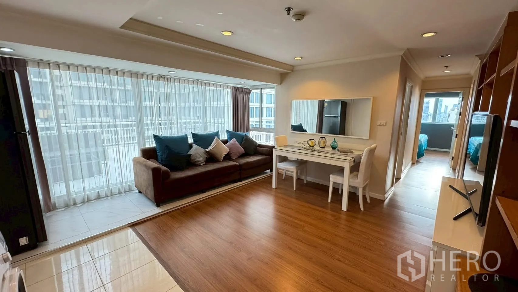 Condominium for rent or sale in Watthana, Bangkok - Bright living room with brown sofa, full-height windows, dining table and hallway leading to bedrooms.