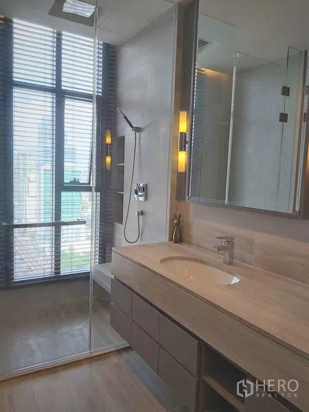Condominium for sale in Bang Rak, Bangkok - Modern bathroom featuring a glass shower, large vanity with mirror cabinets, warm lighting, and a city-facing window.