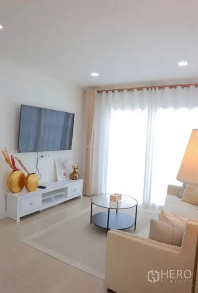 Detached House for rent in Bang Phli, Samut Prakan - Living room with wall-mounted TV, round glass coffee table and sheer curtains.