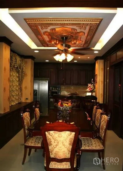 Detached House for sale in Phra Samut Chedi, Samut Prakan - Formal dining room with ornate wood ceiling and fan, upholstered chairs and a built‑in kitchen at the rear.