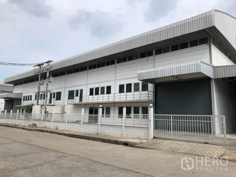Factory for rent in Lam Luk Ka, Pathum Thani - Modern two‑storey factory exterior with metal cladding, fenced frontage and a large roller shutter on a quiet road in Lam Luk Ka.