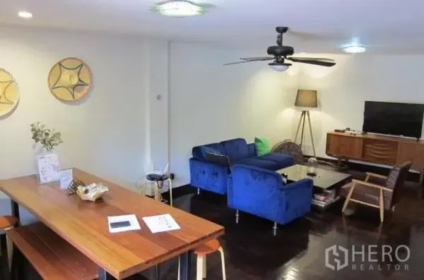 Townhouse for rent in Khlong Toei, Bangkok - Open-plan living and dining room with blue sofa, ceiling fan, wooden table and TV console.