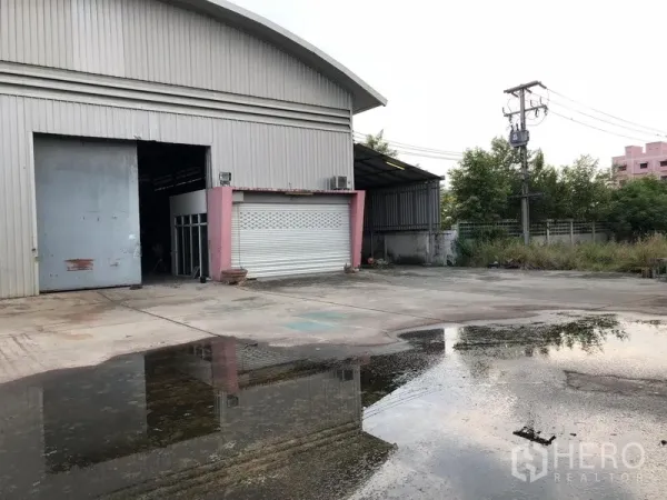 คลังสินค้า ให้เช่าหรือขายในLam Luk Ka, Pathum Thani - โกดังผนังเมทัลชีทพร้อมประตูม้วนขนาดใหญ่ ลานคอนกรีตกว้าง มีรั้วรอบและท่อระบายน้ำ รายล้อมด้วยต้นไม้