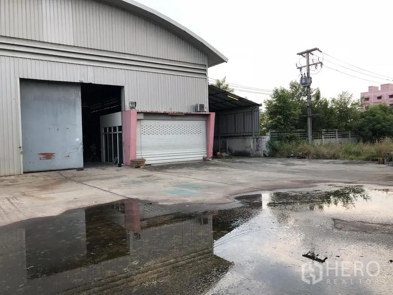คลังสินค้า ให้เช่าหรือขายในLam Luk Ka, Pathum Thani - โกดังผนังเมทัลชีทพร้อมประตูม้วนขนาดใหญ่ ลานคอนกรีตกว้าง มีรั้วรอบและท่อระบายน้ำ รายล้อมด้วยต้นไม้