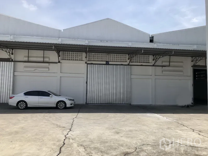 Warehouse for rent in Mueang Pathum Thani, Pathum Thani - Front of white warehouse with large metal sliding doors under an awning and a car parked outside.