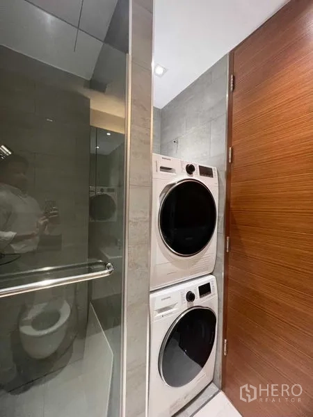 公寓 出售在Khlong Toei, Bangkok - 洗衣区，叠放式洗烘一体机，旁有玻璃浴室门。