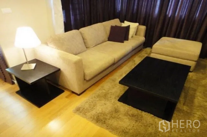 公寓 出租在Sathon, Bangkok - 温馨客厅，米色沙发、脚凳、长绒地毯与深色茶几，配落地窗帘的曼谷公寓。