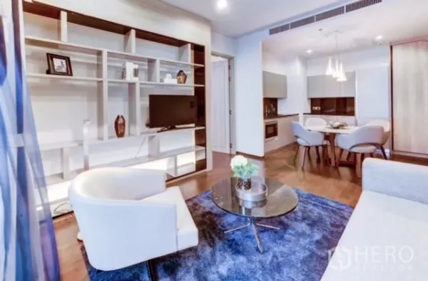 คอนโด ให้เช่าหรือขายในWatthana, Bangkok - ห้องนั่งเล่นชั้นวางบิลต์อินพร้อมทีวี โต๊ะกาแฟกระจกกลม พรมสีน้ำเงิน มองไปยังส่วนทานอาหารและครัว