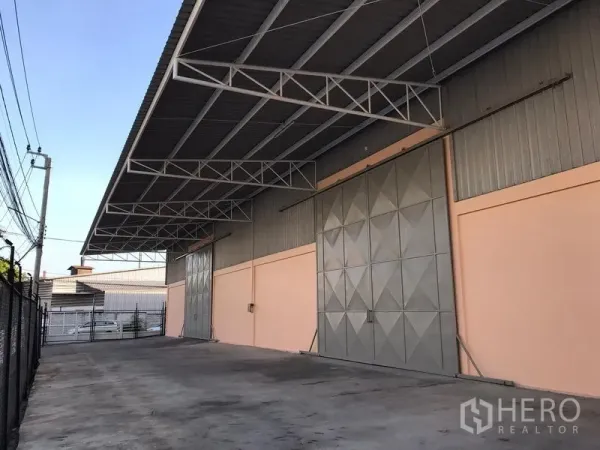 工厂 出租在Mueang Samut Prakan, Samut Prakan - 厂房外立面，带雨棚的装卸区、宽幅推拉钢门和封闭车道。