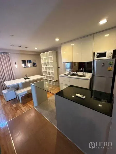 Condominium for sale in Bang Rak, Bangkok - Open-plan kitchen with glossy white cabinets beside dining area and shelving.