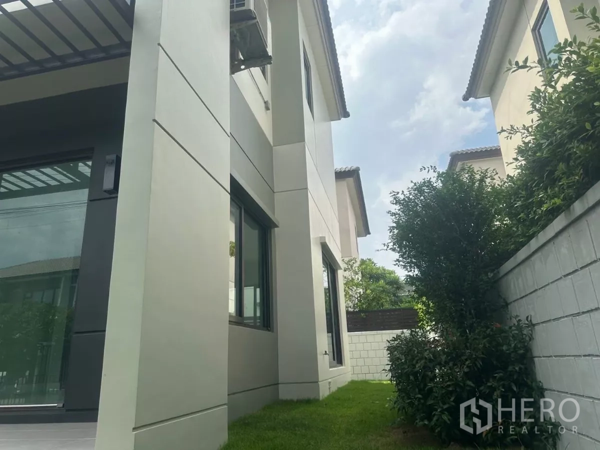Detached House for sale in Samut Prakan - Side view of the house with tall columns, large windows and a green lawn with shrubs along the wall.