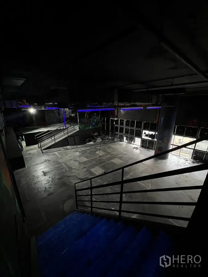 零售空间 出租在Khlong Toei, Bangkok - 带金属栏杆的楼梯通向大型开放式混凝土地面，室内光线较暗。