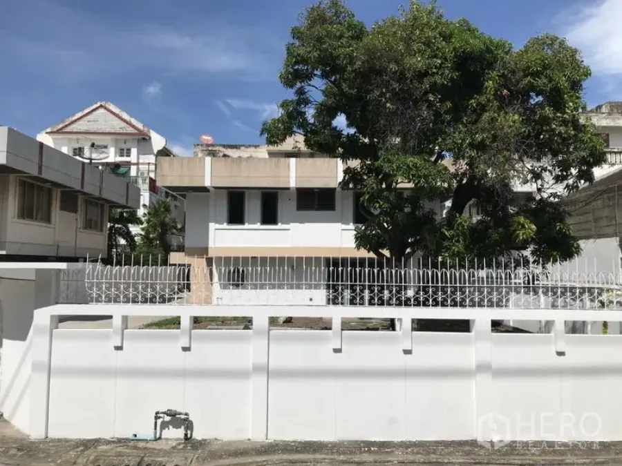 Detached House for rent or sale in Khlong Toei, Bangkok - Two-storey detached house with white perimeter wall, gated driveway and a large shade tree in Khlong Toei under a blue sky.