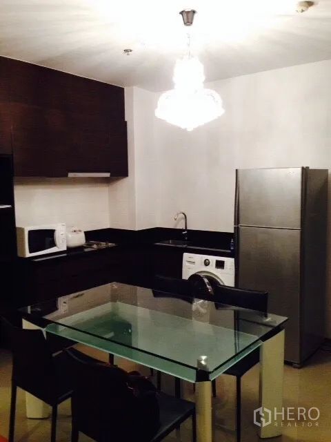 公寓 出租在Phaya Thai, Bangkok - 紧凑厨房，深色橱柜搭配玻璃餐桌与吊灯，配备冰箱、微波炉和洗衣机。