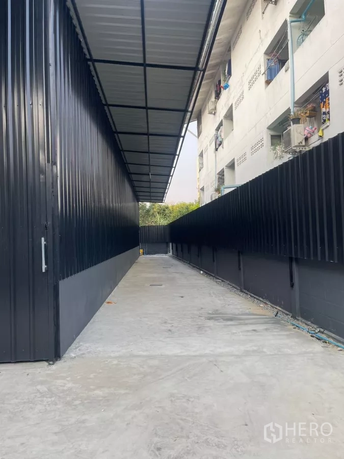 Warehouse for rent in Wang Thonglang, Bangkok - Covered concrete driveway beside a black metal-clad warehouse with secure fencing.