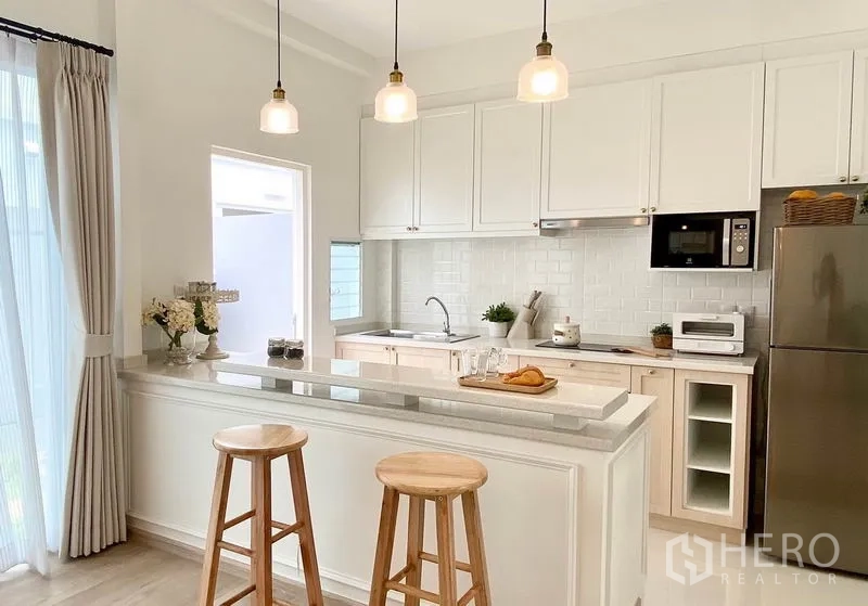 联排别墅 出租在Prawet, Bangkok - 厨房中岛特写，吊灯与吧台凳，白色橱柜