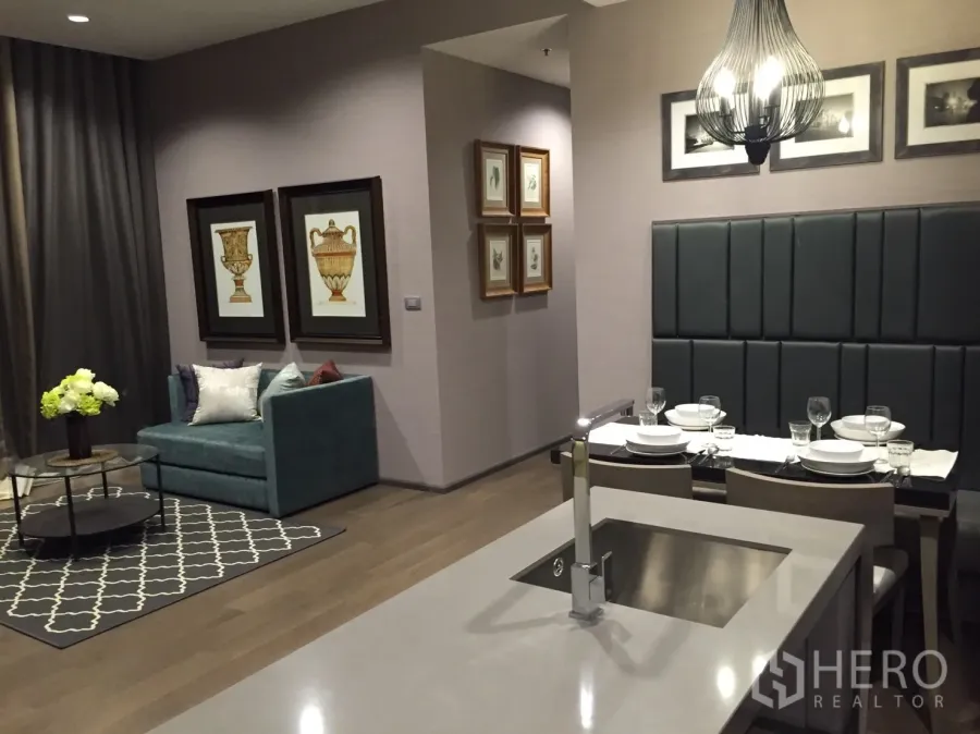 Condominium for rent in Bang Rak, Bangkok - Stylish open-plan living and dining area with teal loveseat, framed art, rug, and kitchen island with sink under a pendant light.