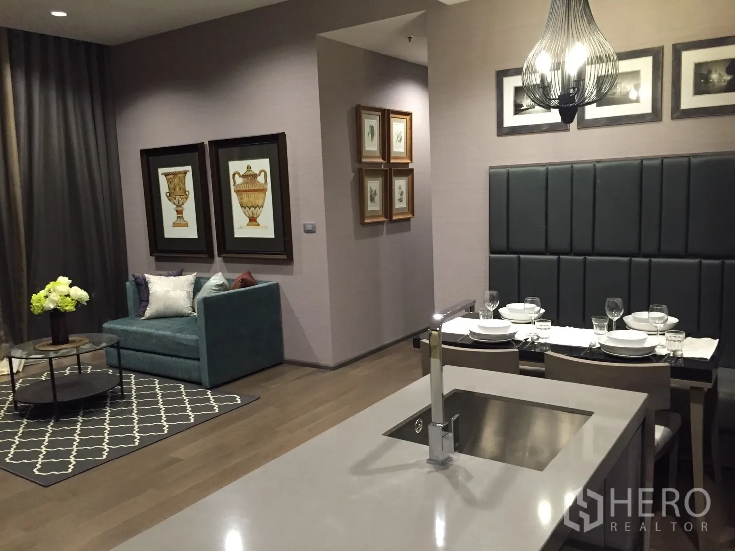 Condominium for rent in Bang Rak, Bangkok - Stylish open-plan living and dining area with teal loveseat, framed art, rug, and kitchen island with sink under a pendant light.