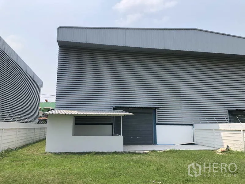 工厂 出售在Sai Noi, Nonthaburi - 工厂后立面，配卷闸门与带遮棚的服务区，旁有草坪。