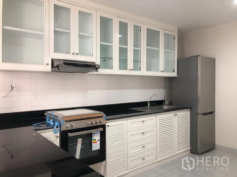 Condominium for rent in Watthana, Bangkok - Bright separate kitchen with white glass-front cabinets, black countertops, subway tile backsplash, oven and stainless refrigerator.