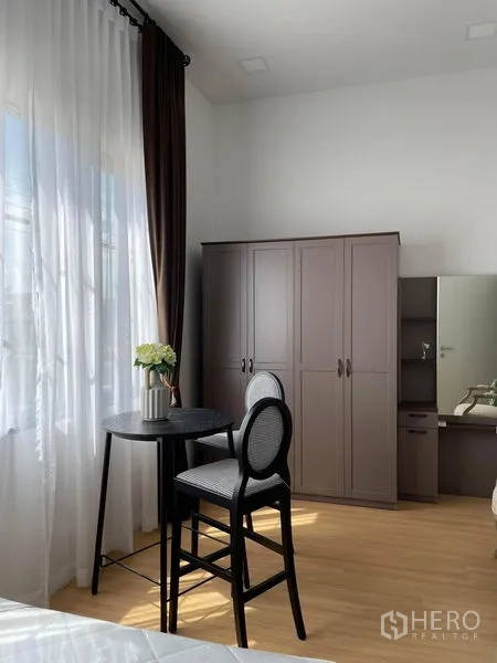 Townhouse for rent in Samut Prakan - Master bedroom corner with tall wardrobe, dressing table and bar-height table by the window.