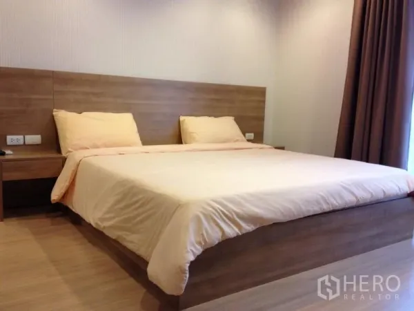 Condominium for rent in Huai Khwang, Bangkok - Spacious primary bedroom with king-size bed, wood accent wall, floor-to-ceiling window, and brown curtains.