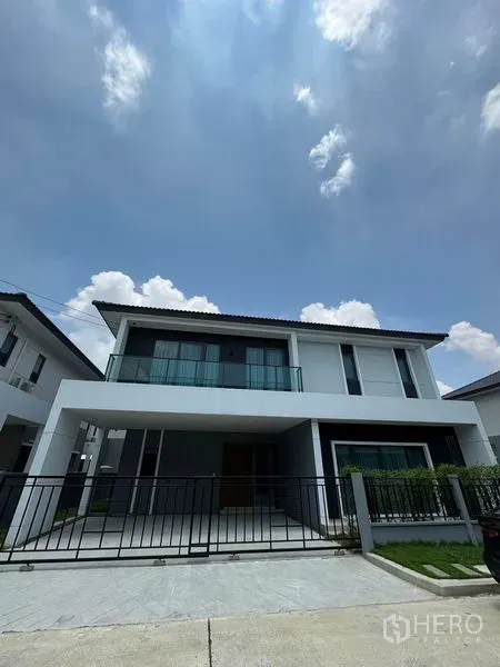 Detached House for rent in Don Mueang, Bangkok - Modern two-storey detached house in Don Mueang with glass balcony, gated driveway and small lawn under a bright sky.