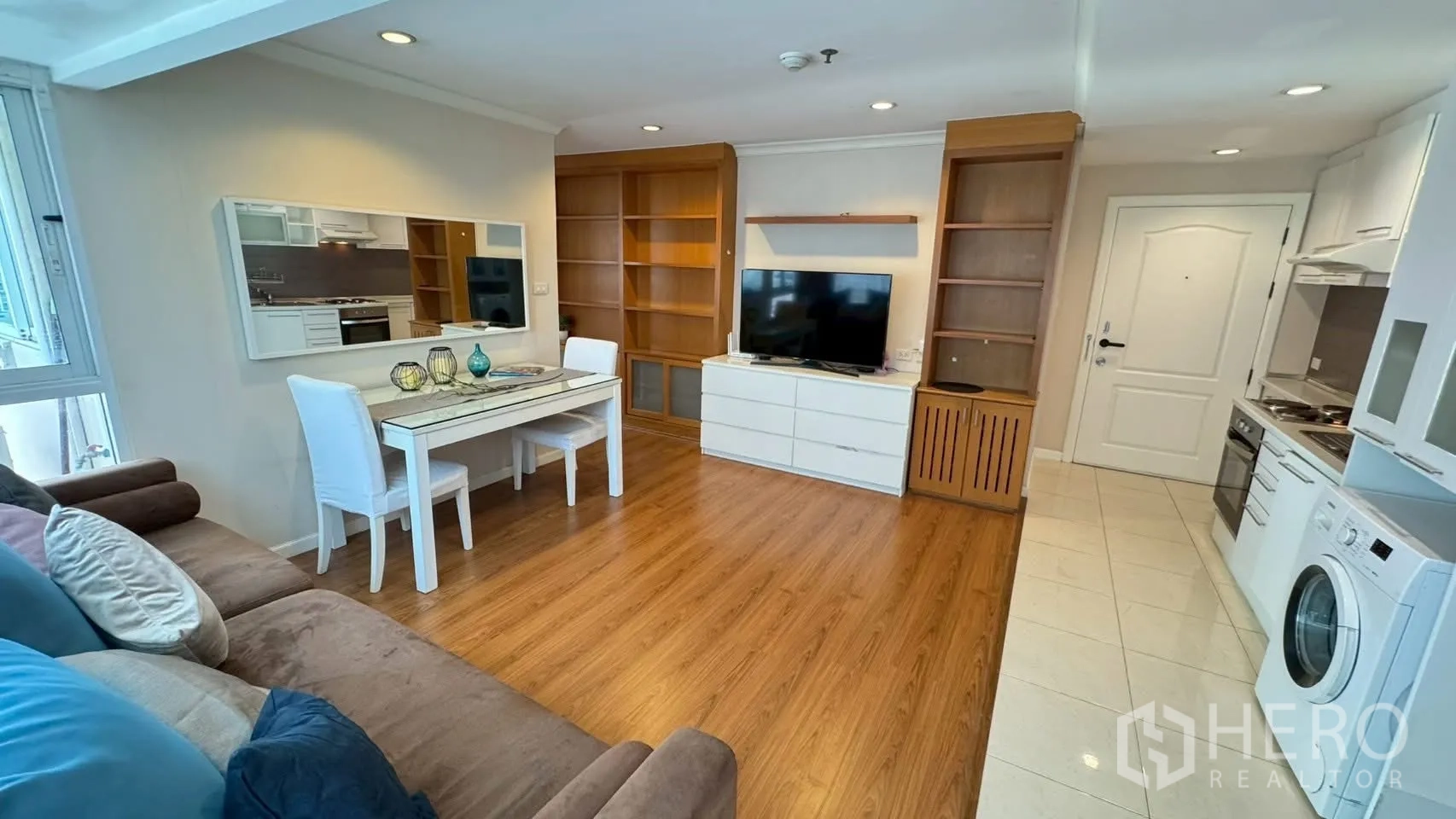 Condominium for rent or sale in Watthana, Bangkok - Living space with TV console, white dining set and built-in wooden shelving on warm wood floors.