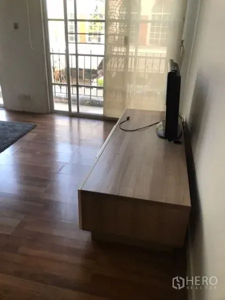 Townhouse for rent in Bang Kapi, Bangkok - Bright living area with wooden TV console, wood floors, and sliding glass doors opening to a small balcony.