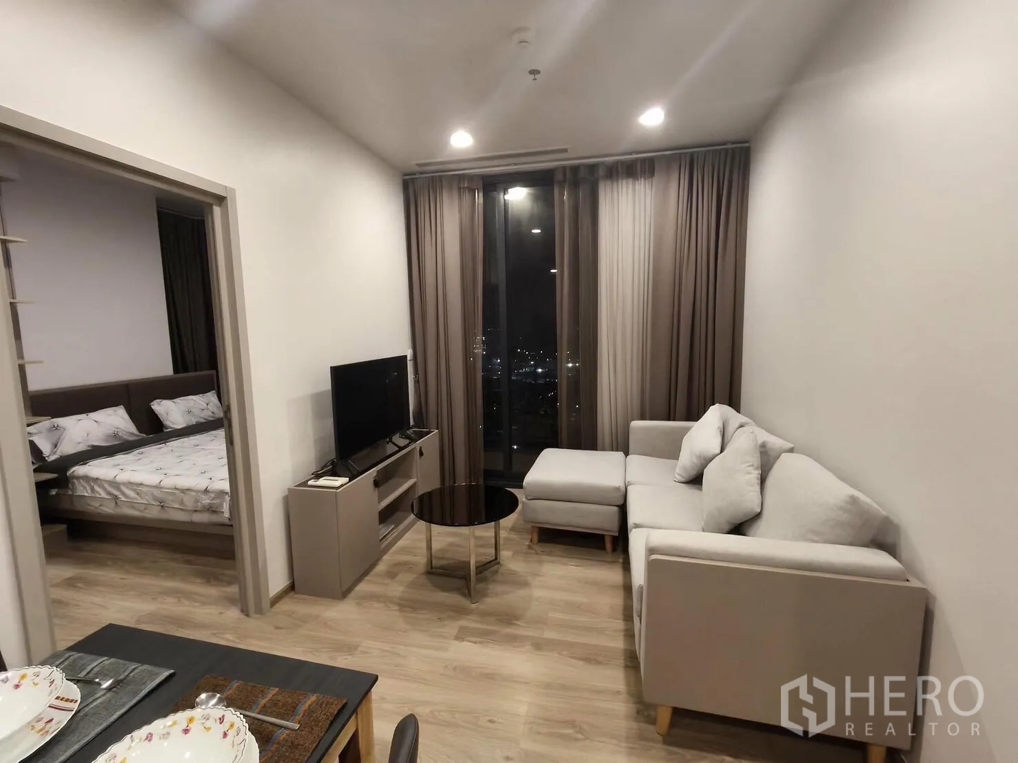 คอนโด for rent in Khlong Toei, Bangkok - ห้องนั่งเล่นอบอุ่นมีโซฟาเข้ามุม ตู้วางทีวี ผ้าม่านสูง และวิวเมือง พร้อมประตูสู่ห้องนอน