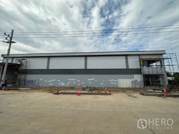 Warehouse for rent or sale in Lam Luk Ka, Pathum Thani - Full‑length facade of the warehouse facing a 16‑meter concrete road with safety cones.
