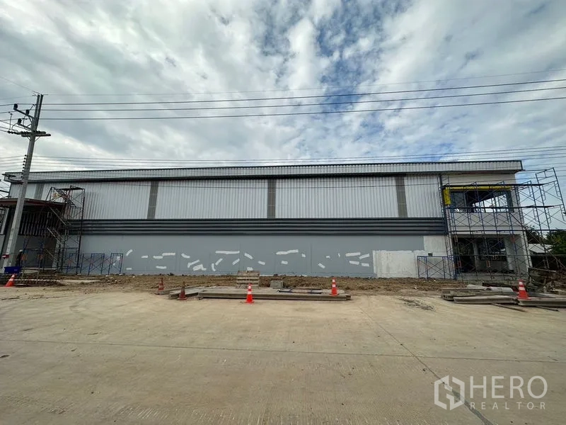 Warehouse for rent or sale in Lam Luk Ka, Pathum Thani - Full‑length facade of the warehouse facing a 16‑meter concrete road with safety cones.