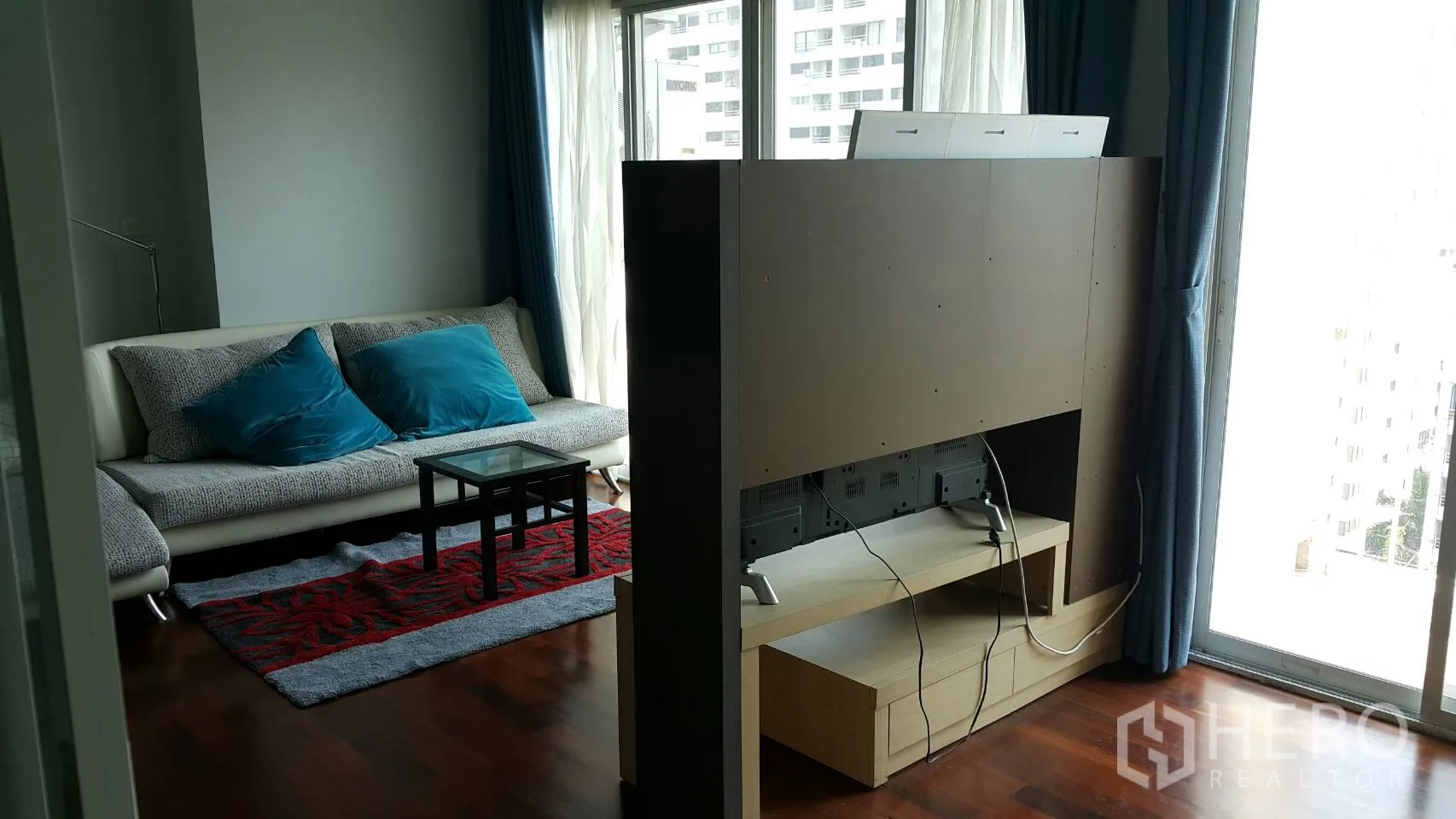 Condominium for rent in Watthana, Bangkok - Lounge with TV partition, large windows, blue curtains and city views beyond the balcony.