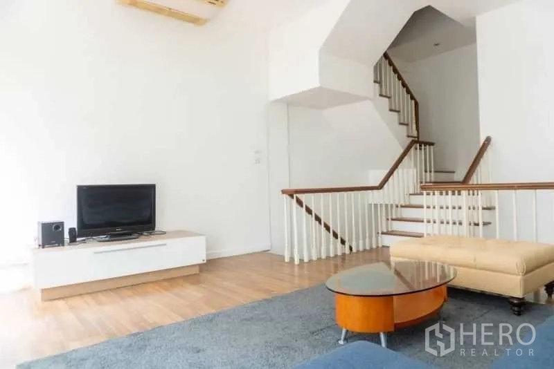 Townhouse for rent in Bang Kapi, Bangkok - Living room with TV unit, coffee table, sectional sofa, blue rug, and white-railed staircase.