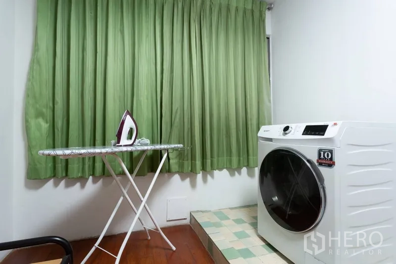 Condominium for rent or sale in Ratchathewi, Bangkok - Compact laundry nook with front-load washing machine, ironing board and green curtains.