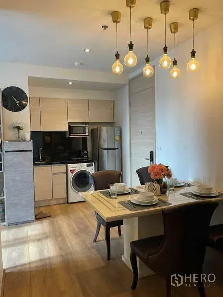 Condominium for rent or sale - Open-plan kitchen with light wood cabinets, black countertop, fridge, microwave and washing machine beside a dining table under warm pendant lights.