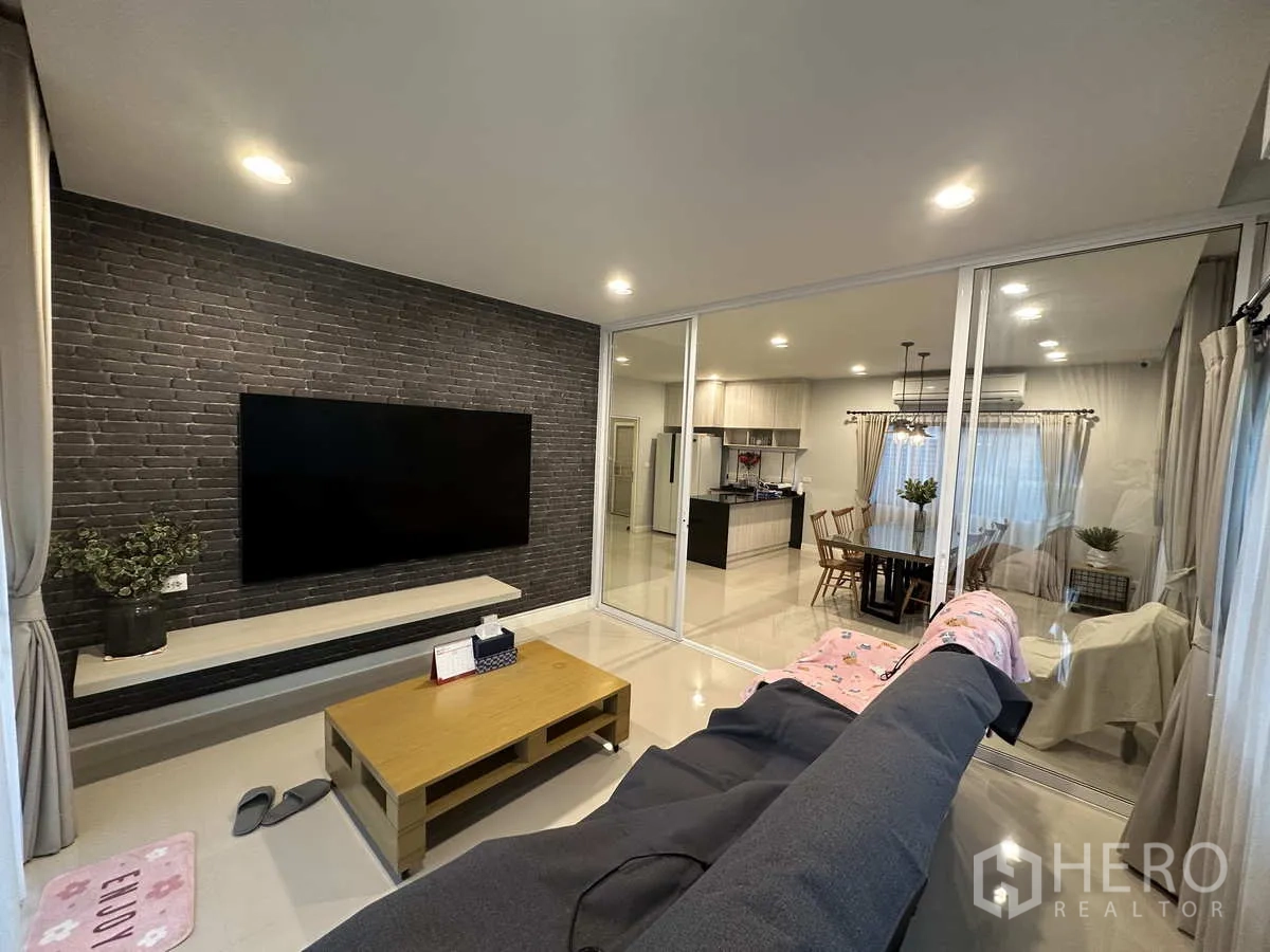 บ้าน ให้เช่าหรือขายในLam Luk Ka, Pathum Thani - ห้องนั่งเล่นอบอุ่น ผนังก่ออิฐตกแต่ง ทีวีจอใหญ่ และประตูบานเลื่อนกระจก.