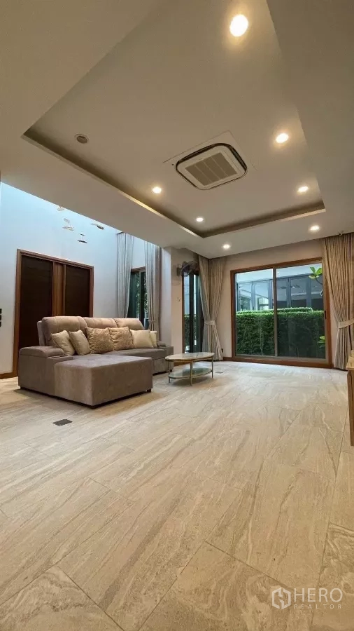 Detached House for rent in Prawet, Bangkok - Living area with large sliding glass doors opening to a hedge-lined garden and an L-shaped sofa.