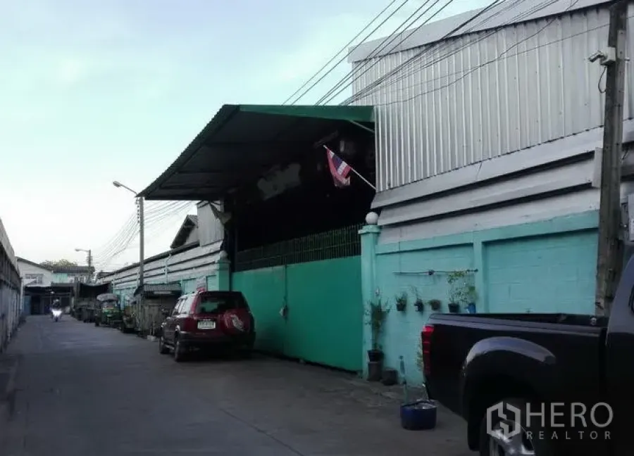 คลังสินค้า ให้เช่าในBang Kho Laem, Bangkok - แถวโกดังโครงสร้างเมทัลชีท ผนังสีเขียว มีหลังคาคลุมลานขนถ่าย ธงชาติไทย และตรอกทางเข้า-ออกที่มีรถจอด ในย่านบางคอแหลม