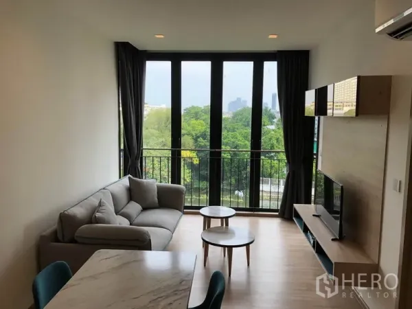 คอนโด ให้เช่าในWatthana, Bangkok - ห้องนั่งเล่นโปร่งที่ Kawa Haus มีหน้าต่างสูงจรดเพดาน ระเบียง และวิวต้นไม้ พร้อมโซฟา ทีวี และโต๊ะกลาง