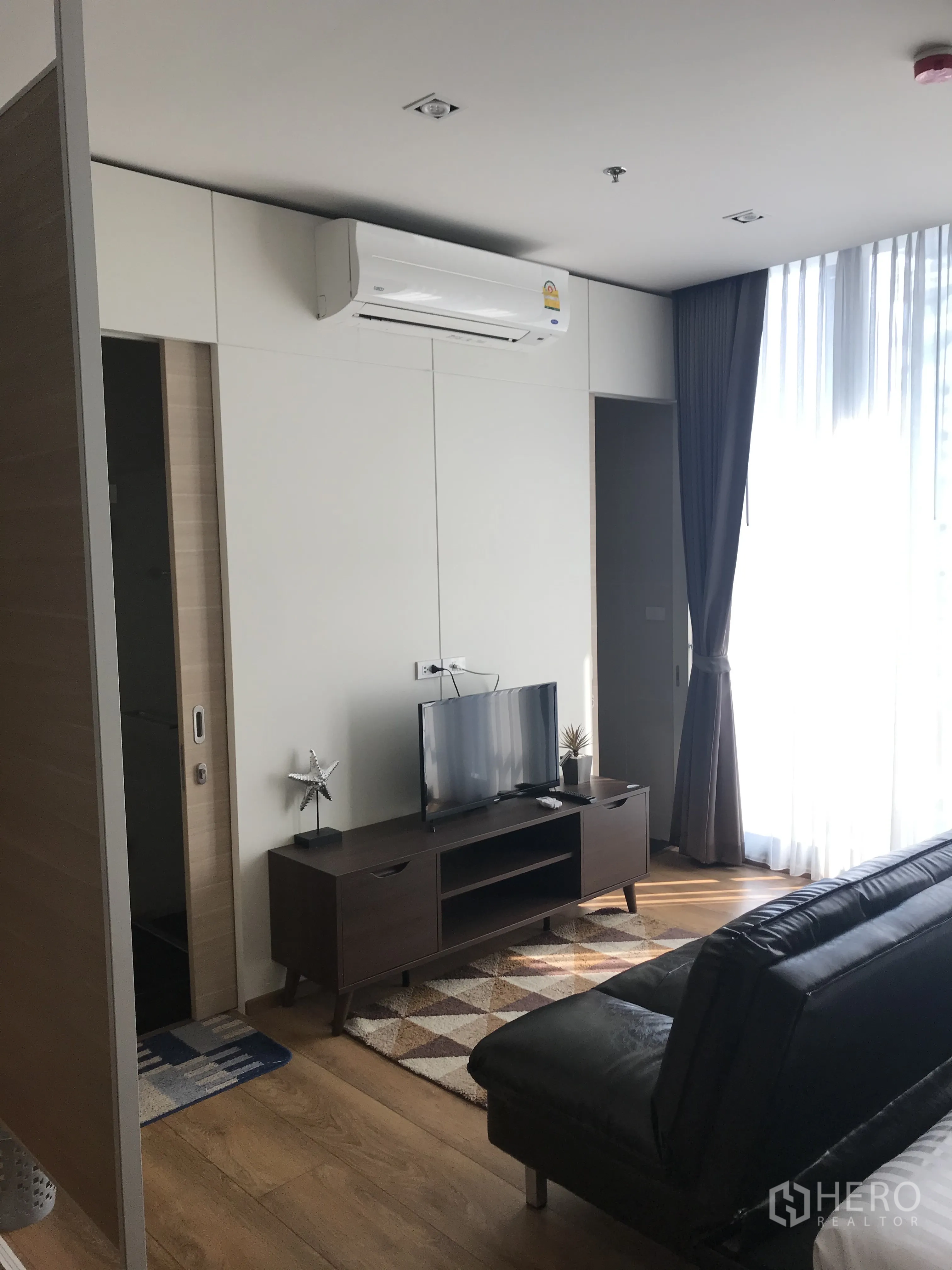 Condominium for sale in Khlong Toei, Bangkok - Living area with TV console, rug, leather sofa, and large curtained windows.