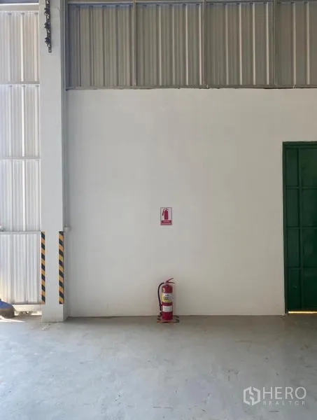 Warehouse for rent in Mueang Rayong, Rayong - Safety view of a fire extinguisher mounted on a white wall beside a steel sliding door.