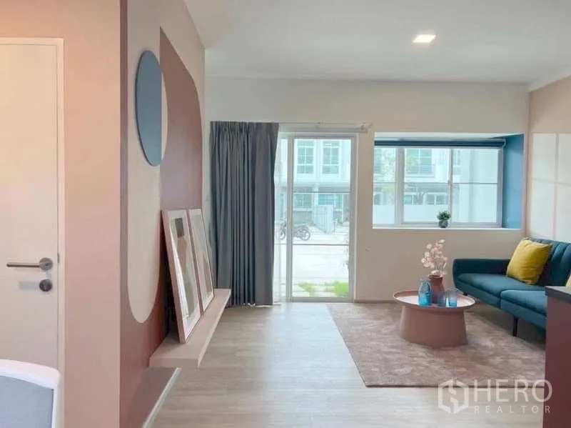 Townhouse for rent in Bang Bo, Samut Prakan - Open-plan living area with sliding glass door, big window, soft rug, blue sofa and pastel coffee table.