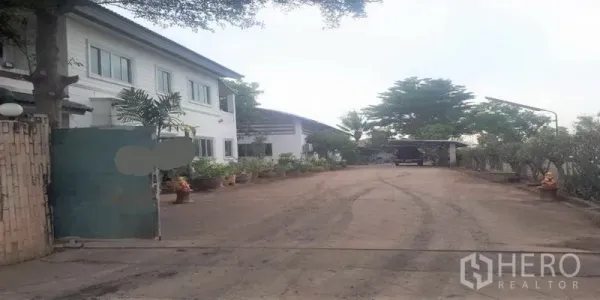 货栈 出售在Pathum Thani - 带大门的入口与宽阔混凝土车道，通向仓库和办公楼，摆放花盆。