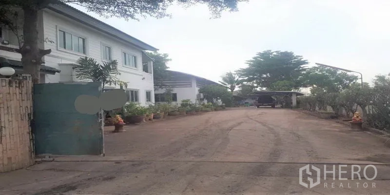 货栈 出售在Pathum Thani - 带大门的入口与宽阔混凝土车道，通向仓库和办公楼，摆放花盆。